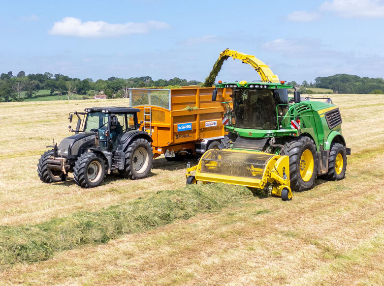 Valtra tractor towing a SW Hire 14t Richard Western Silage Trailer, filling up with grass from a John Deere Forager in a grass stubble field with blue skies